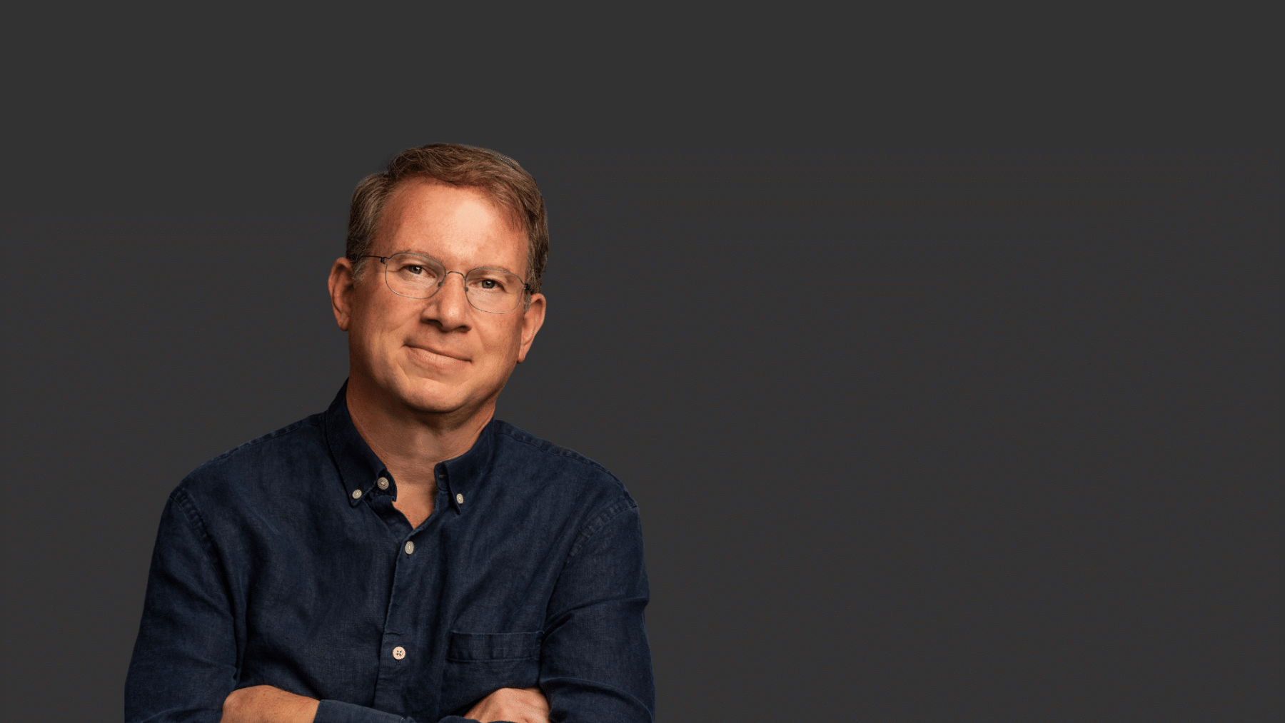 Jeffrey Goldberg, a light skinned man with short brown hair stands with arms folded wearing a navy blue button down shirt.